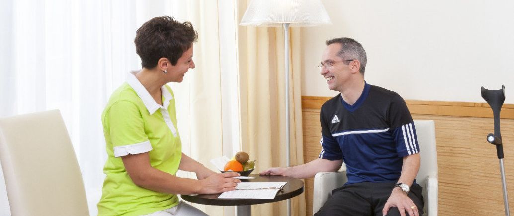 Mitarbeiterin Roomservice bespricht sich mit Patient am Tisch